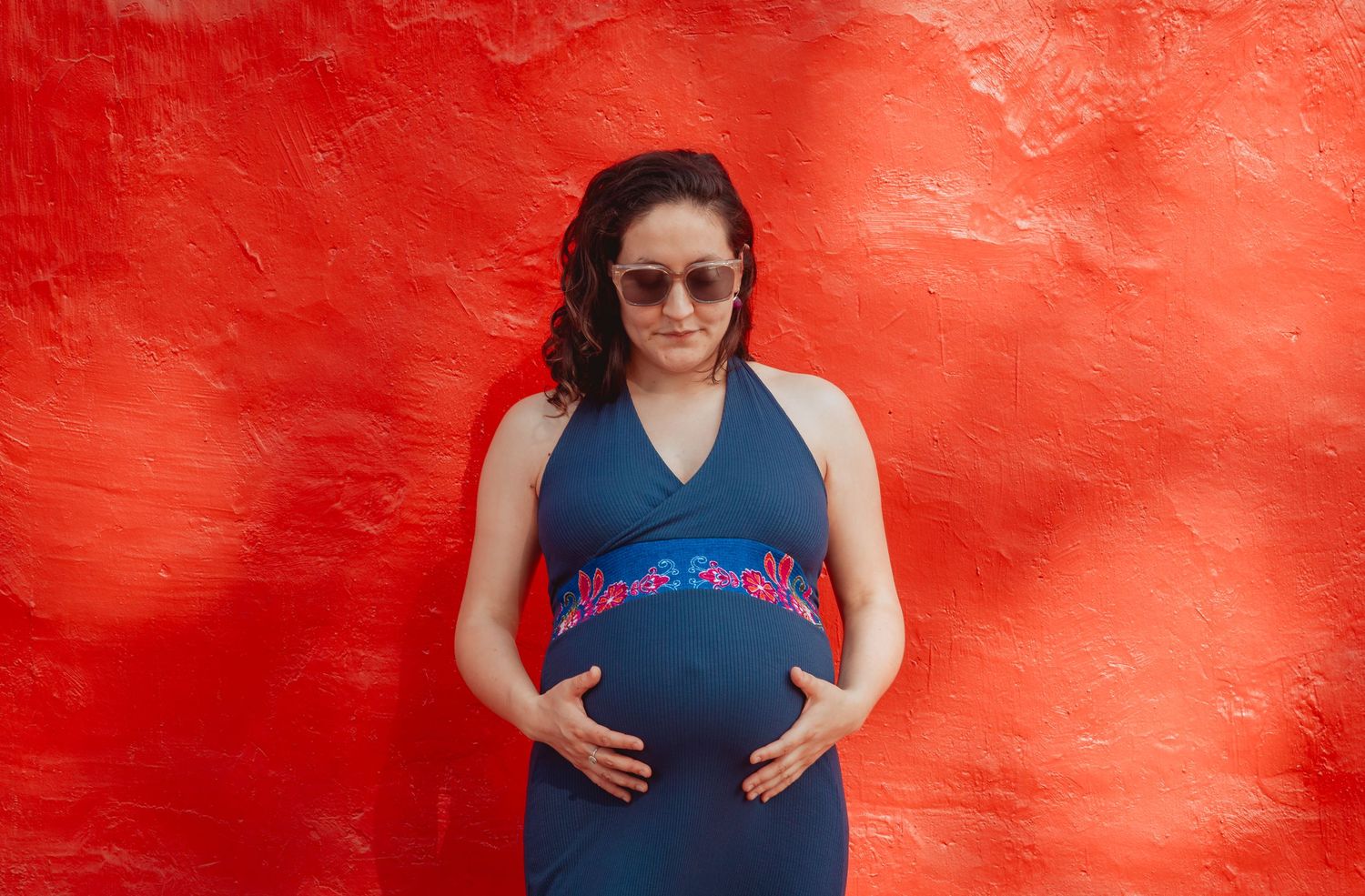Pregnant woman in sunglasses cradling her belly against a vivid red wall.