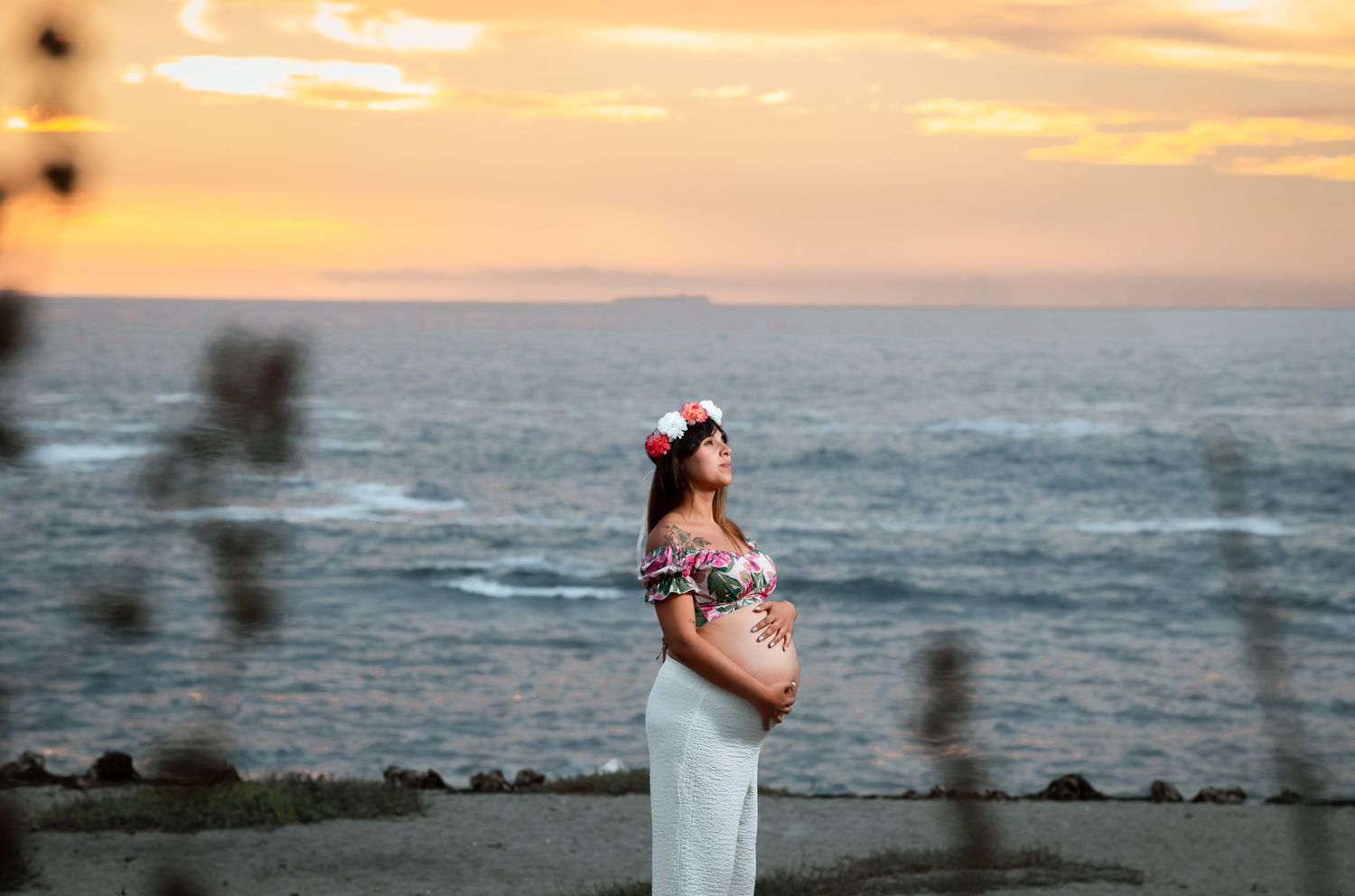 Pregnant woman in floral top and white skirt poses gracefully by the ocean at sunset.