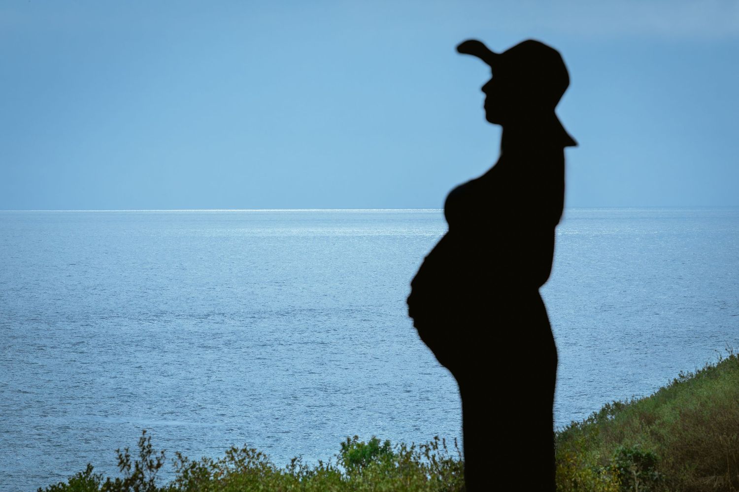 Silhouette of pregnant woman standing by ocean wearing cap.