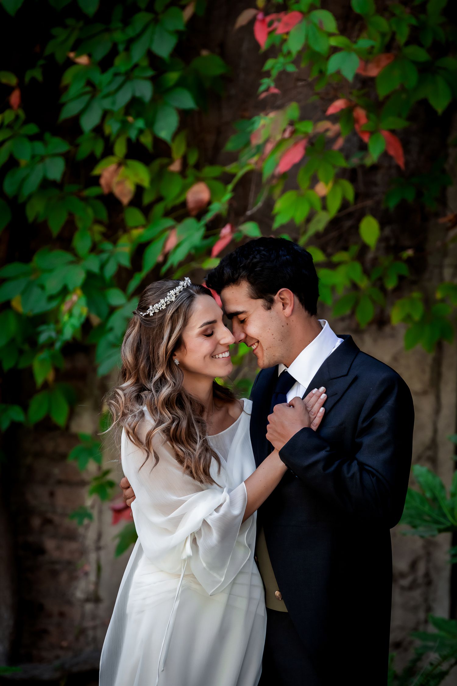 Bride wearing a tiara and groom in a black tuxedo share a romantic embrace before colorful foliage.