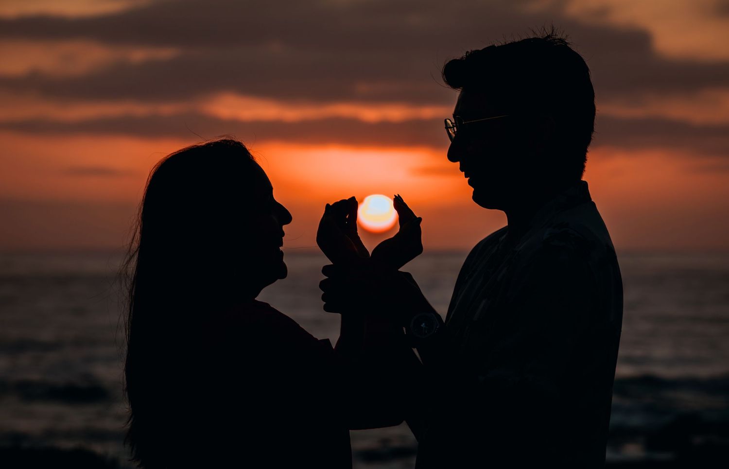 Romantic couple silhouetted against a vibrant ocean sunset, holding a wine glass capturing the glowing sun.