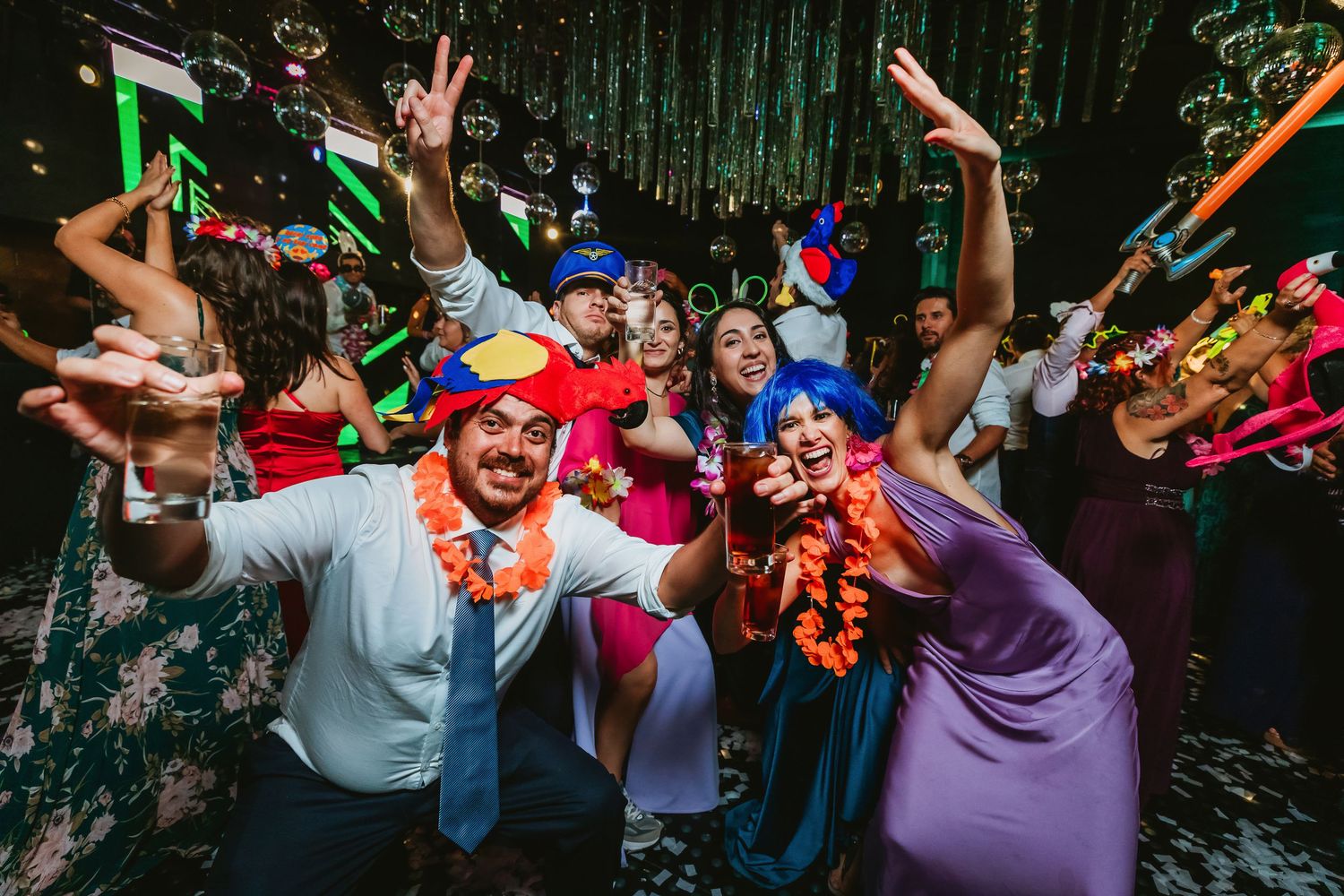 Festive party guests wearing colorful wigs and accessories celebrating on a dance floor at night.