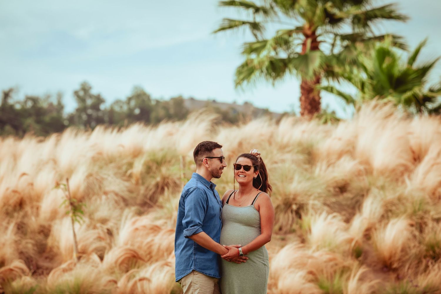 Smiling pregnant couple standing together among swaying golden pampas grass under a partly cloudy sky.