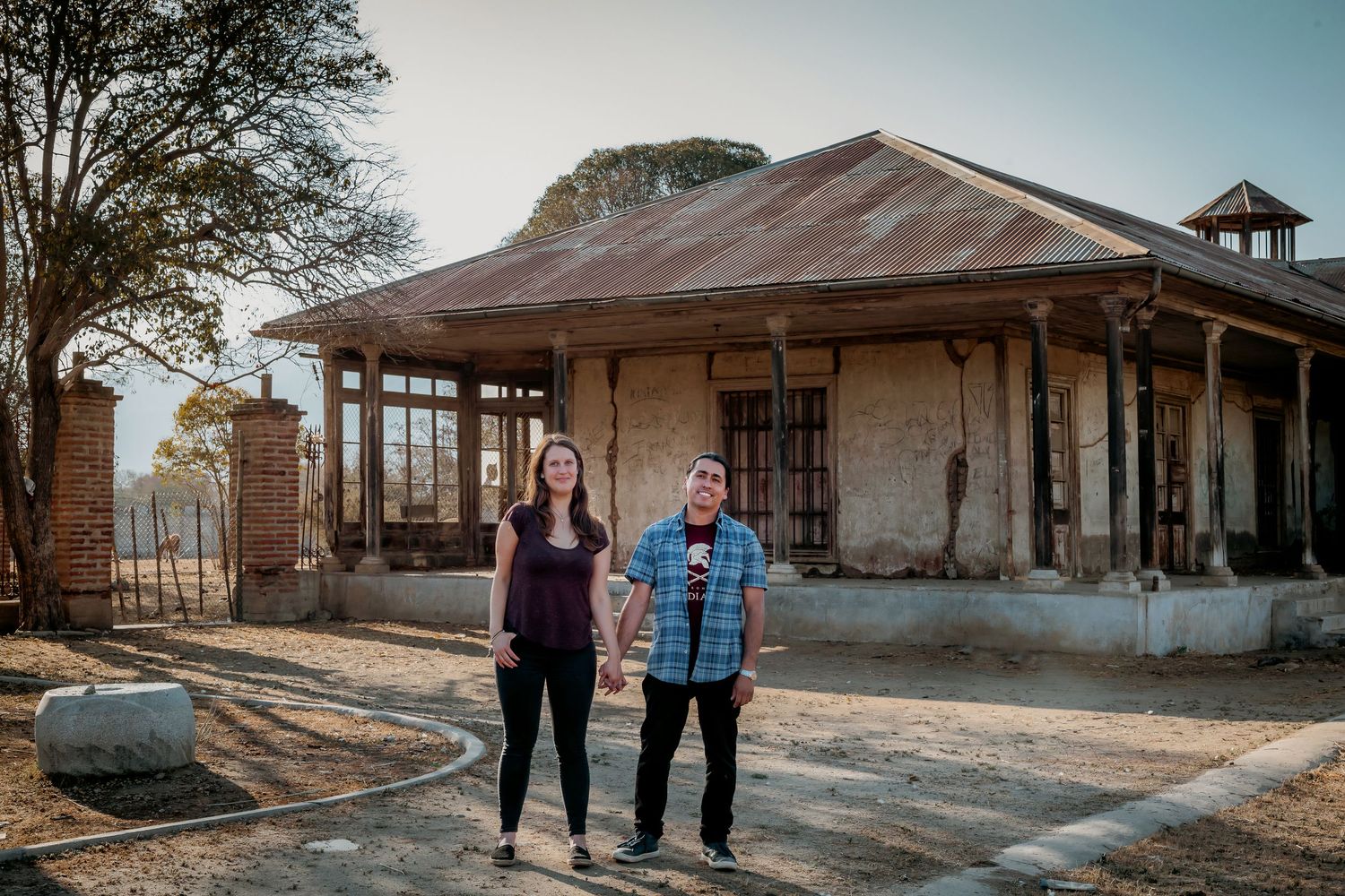 A couple holds hands standing in front of a rustic abandoned building with a corrugated metal roof.