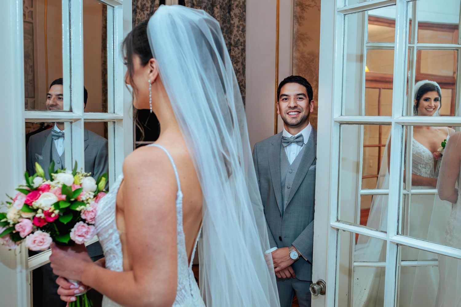 Groom smiles at bride in white wedding dress and veil during first look moment at elegant venue.