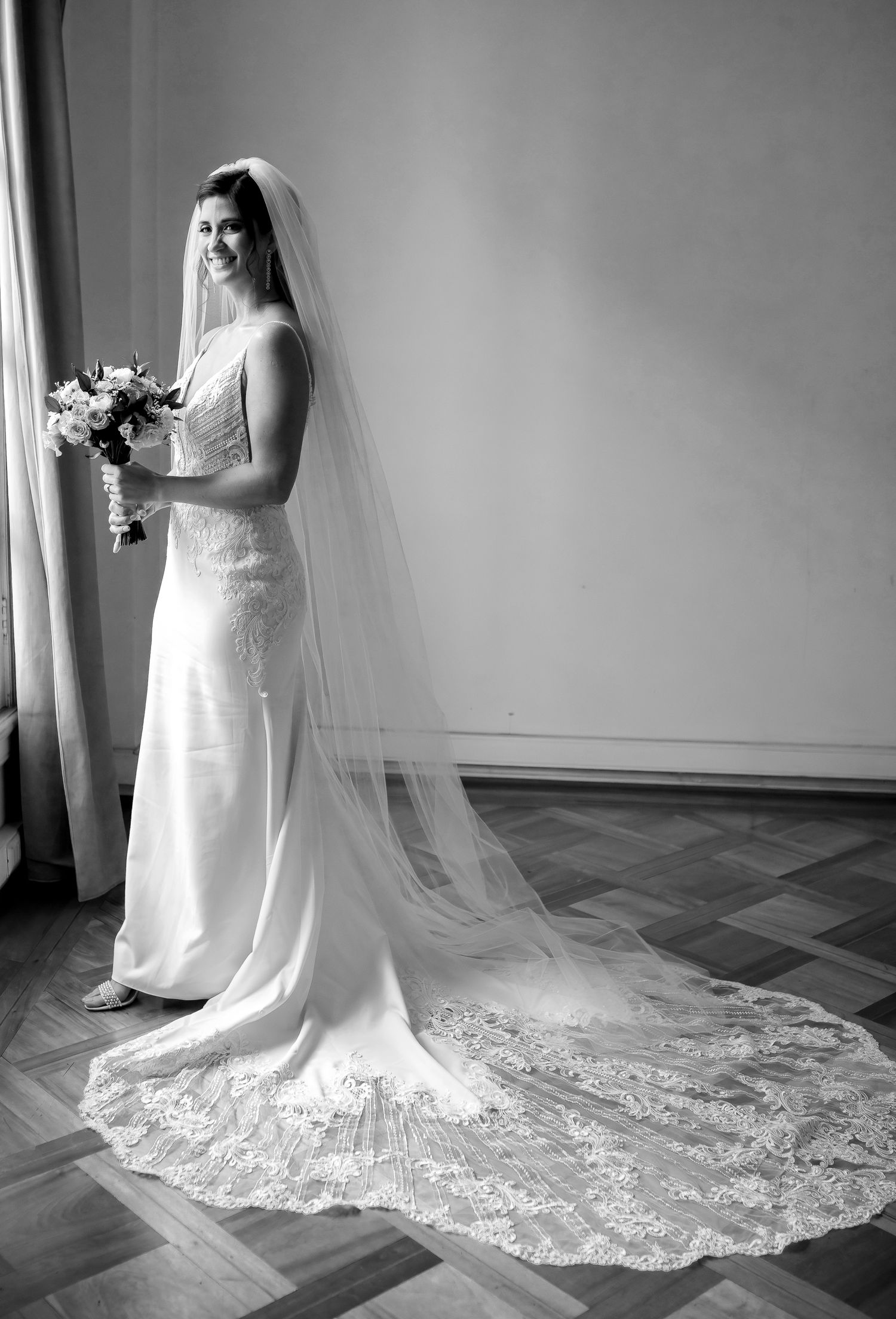 Bride in elegant lace wedding gown with long cathedral veil holding bouquet, black and white portrait photo.