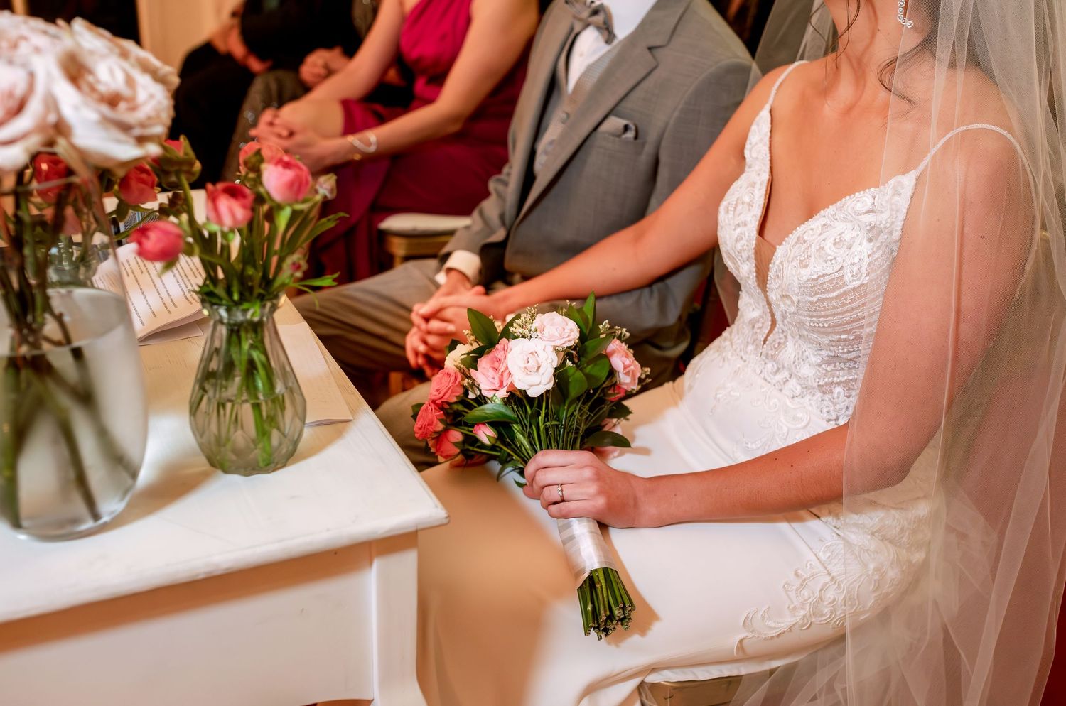 Bride in white lace gown holding pink bouquet at wedding ceremony with floral arrangements on table.