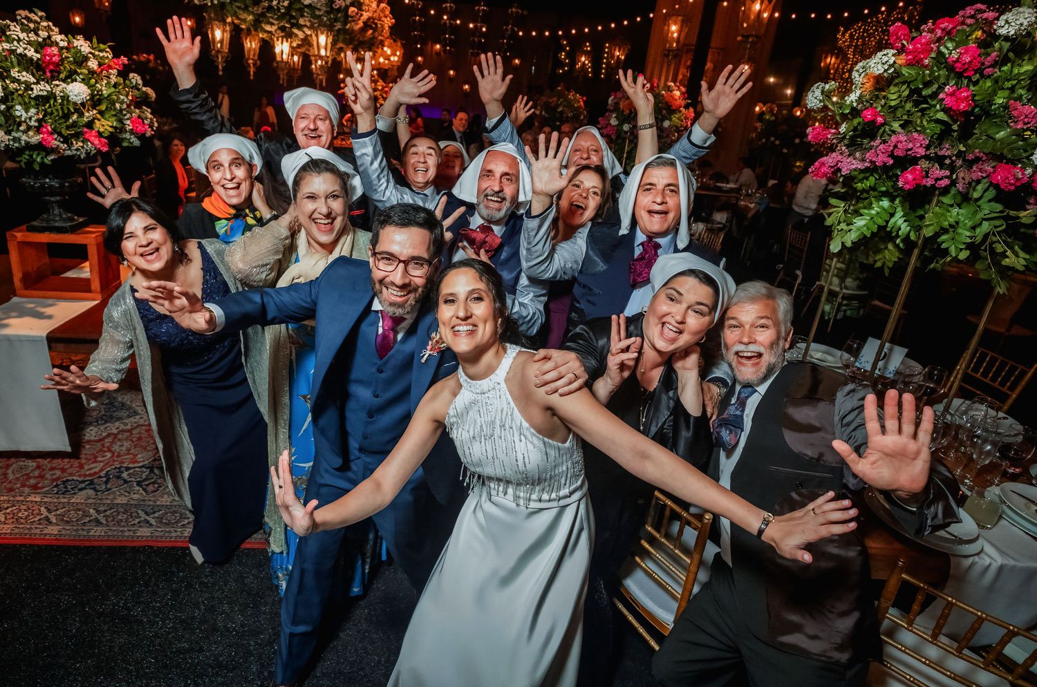 Joyful wedding party group photo with bride and groom surrounded by celebrating guests outdoors.