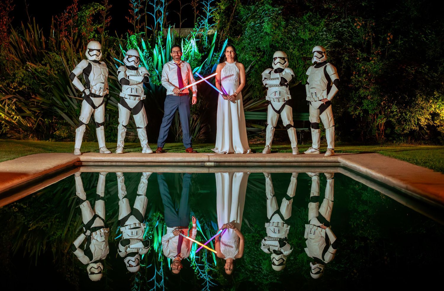 Newlyweds hold crossed lightsabers flanked by Stormtroopers with a stunning pool reflection.