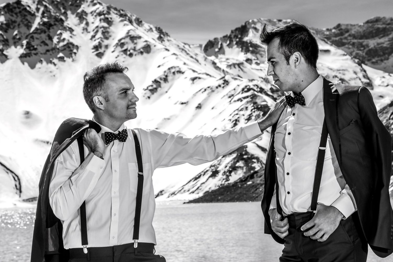 Black and white photo of two grooms in bow ties and suspenders laughing by a snowy mountain lake.