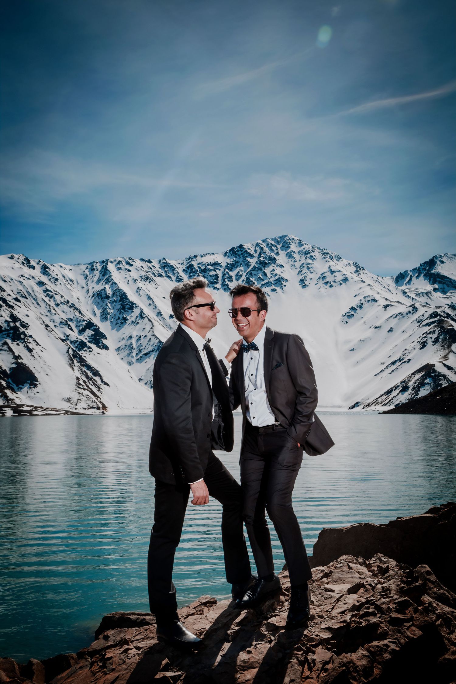 Two grooms in dark suits share a romantic moment by a turquoise alpine lake with snow-capped mountains.