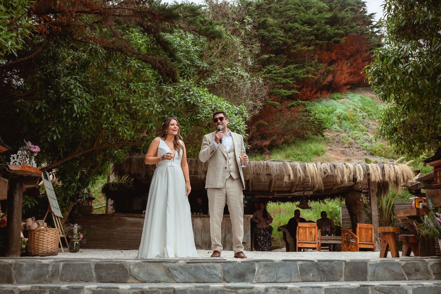 Bride and groom stand together on stone steps at an outdoor wedding venue surrounded by lush green trees.