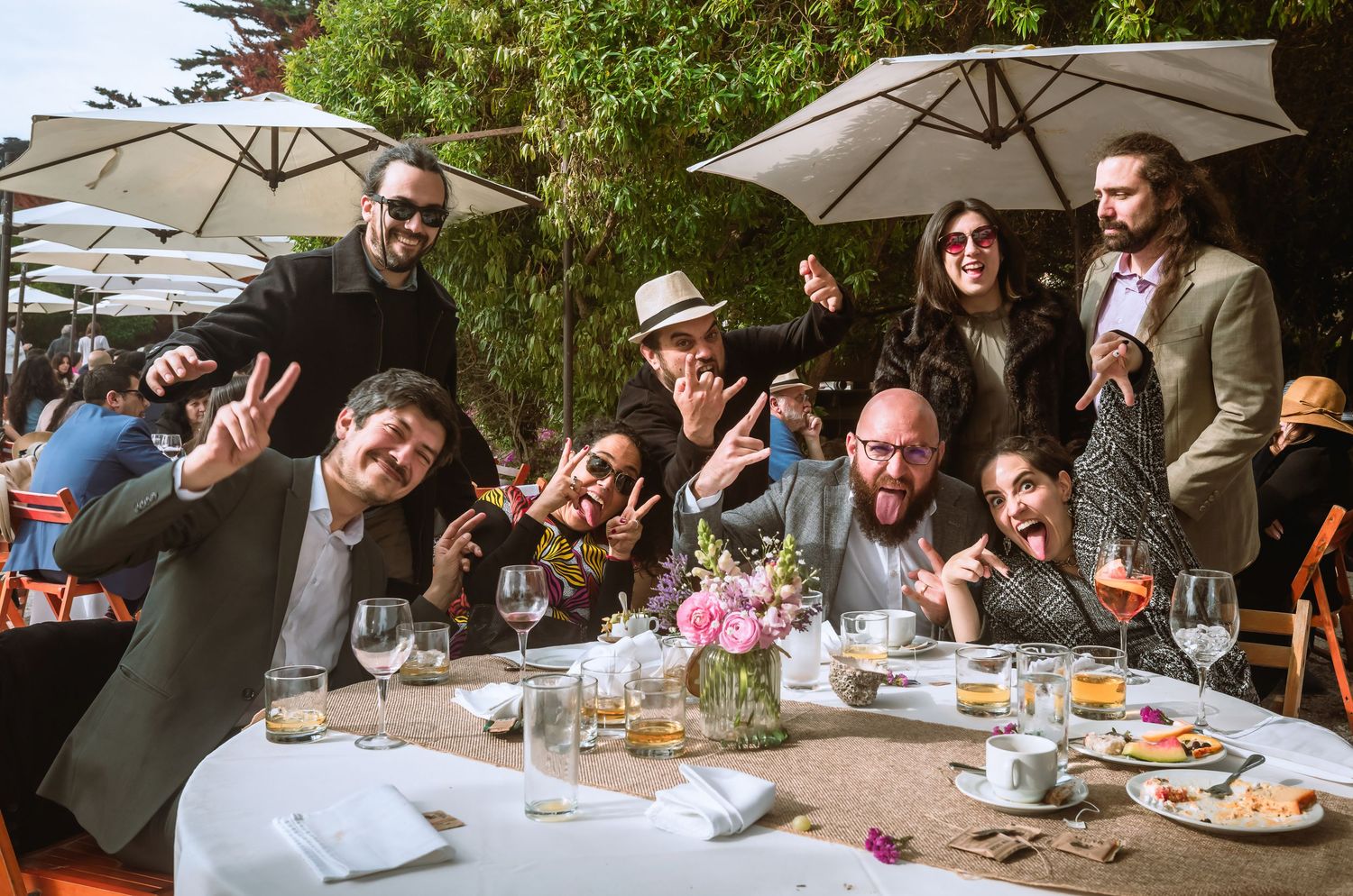 Group of friends posing and making fun gestures at an outdoor wedding reception dinner table with flowers and wine glasses.