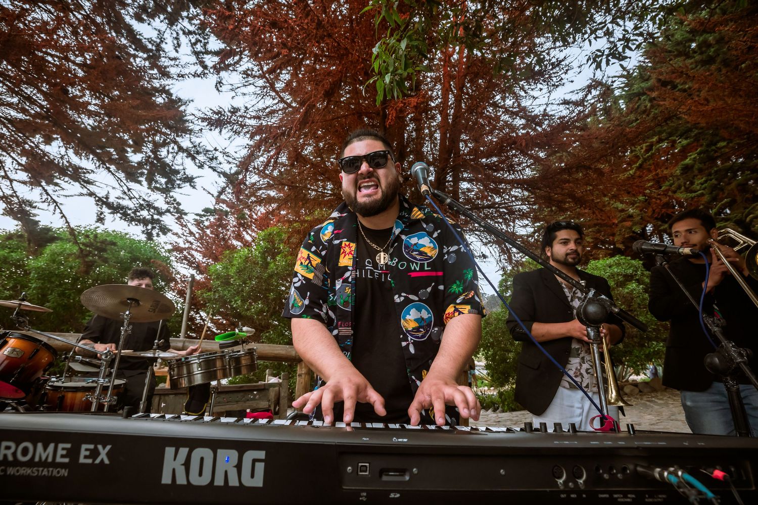 Musician playing a Korg keyboard synthesizer outdoors at a wedding reception under red autumn trees.