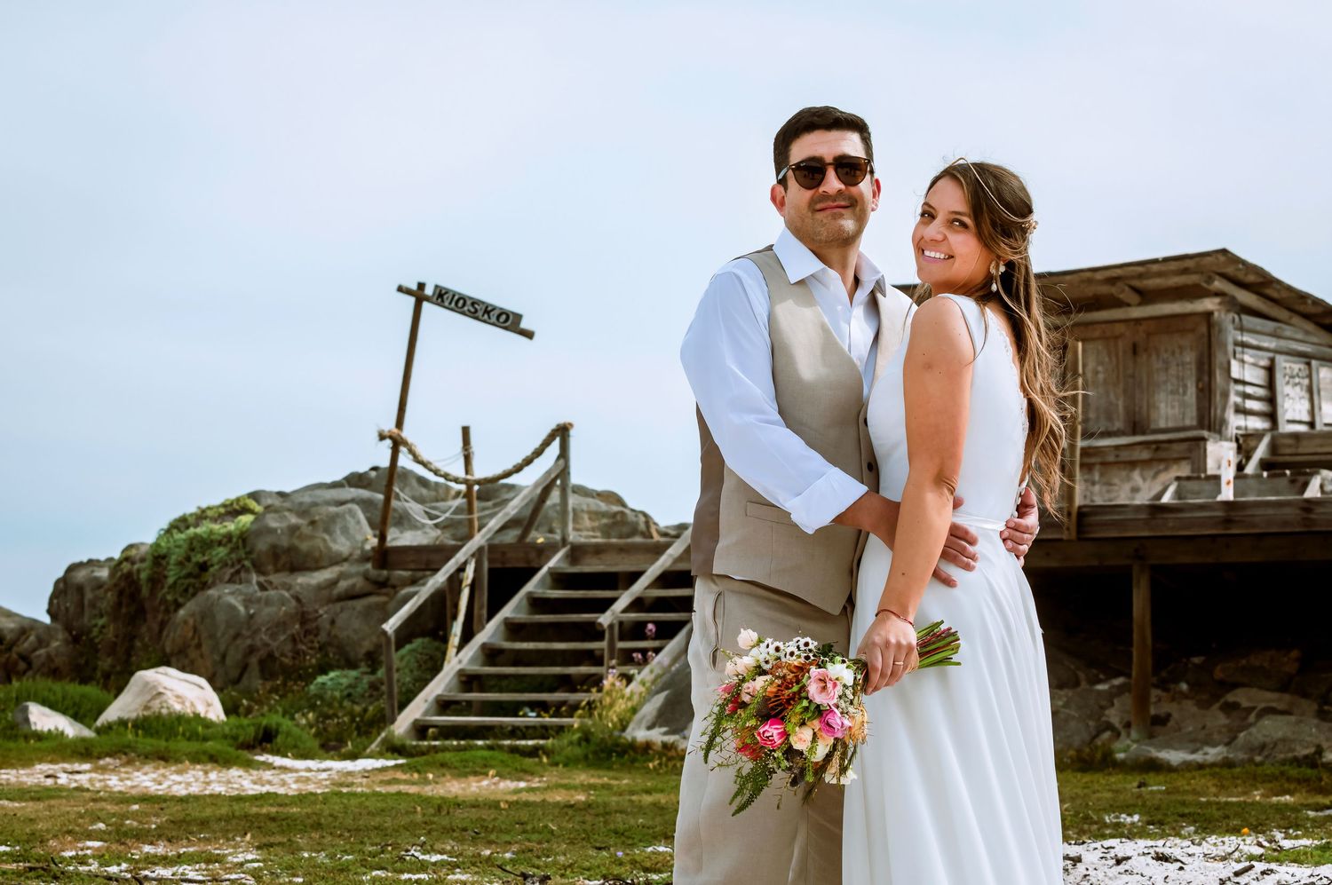 Bride and groom posing together near rustic wooden structure and rocky beach landscape on wedding day.