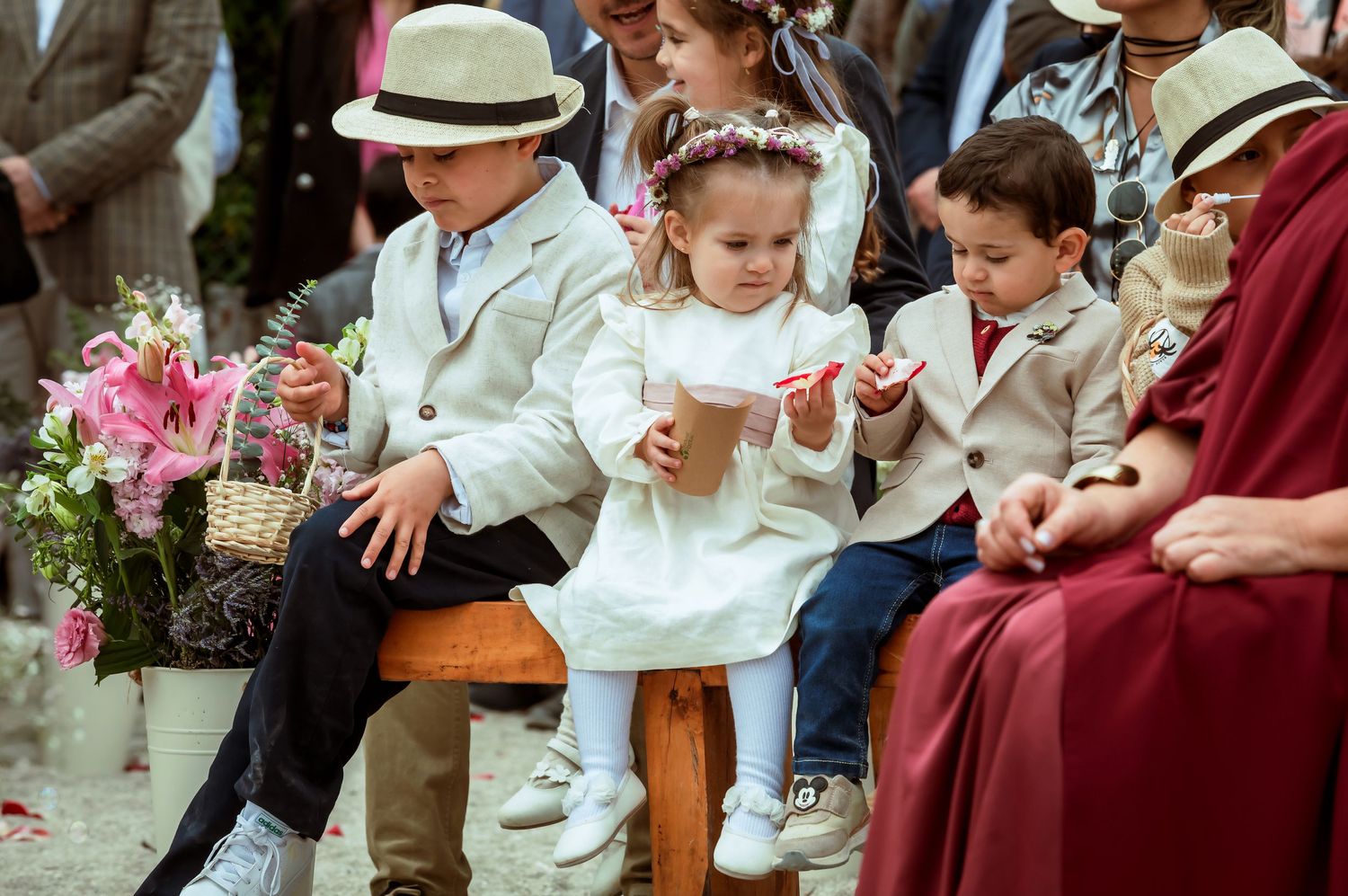 Three young children in white formal attire sit together during an outdoor wedding ceremony.