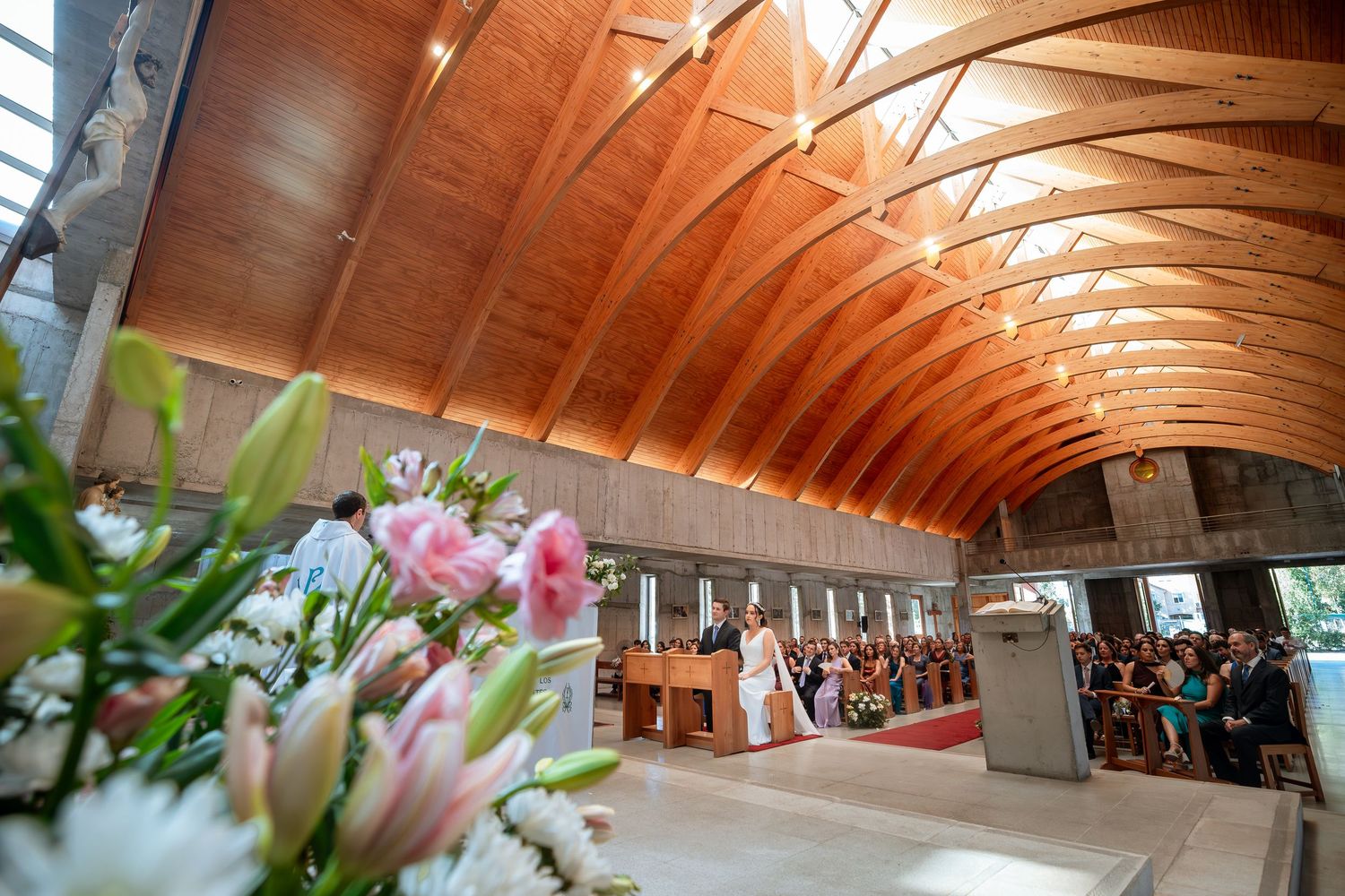 Wide-angle view of church wedding with wooden arched ceiling and floral decorations.