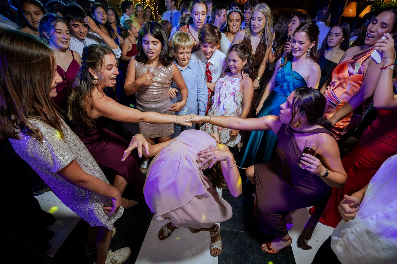 Excited wedding guests forming a circle on the dance floor around a woman in white bending down.