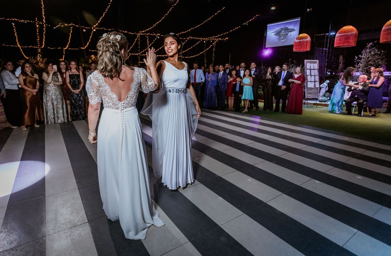 Two brides in light blue gowns share first dance as wedding guests watch in background.