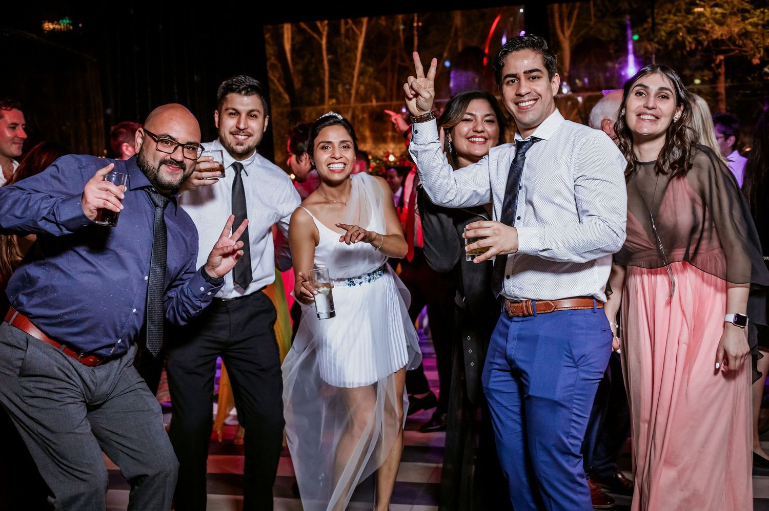 Bride in white dress poses with cheerful wedding guests at outdoor nighttime reception party.