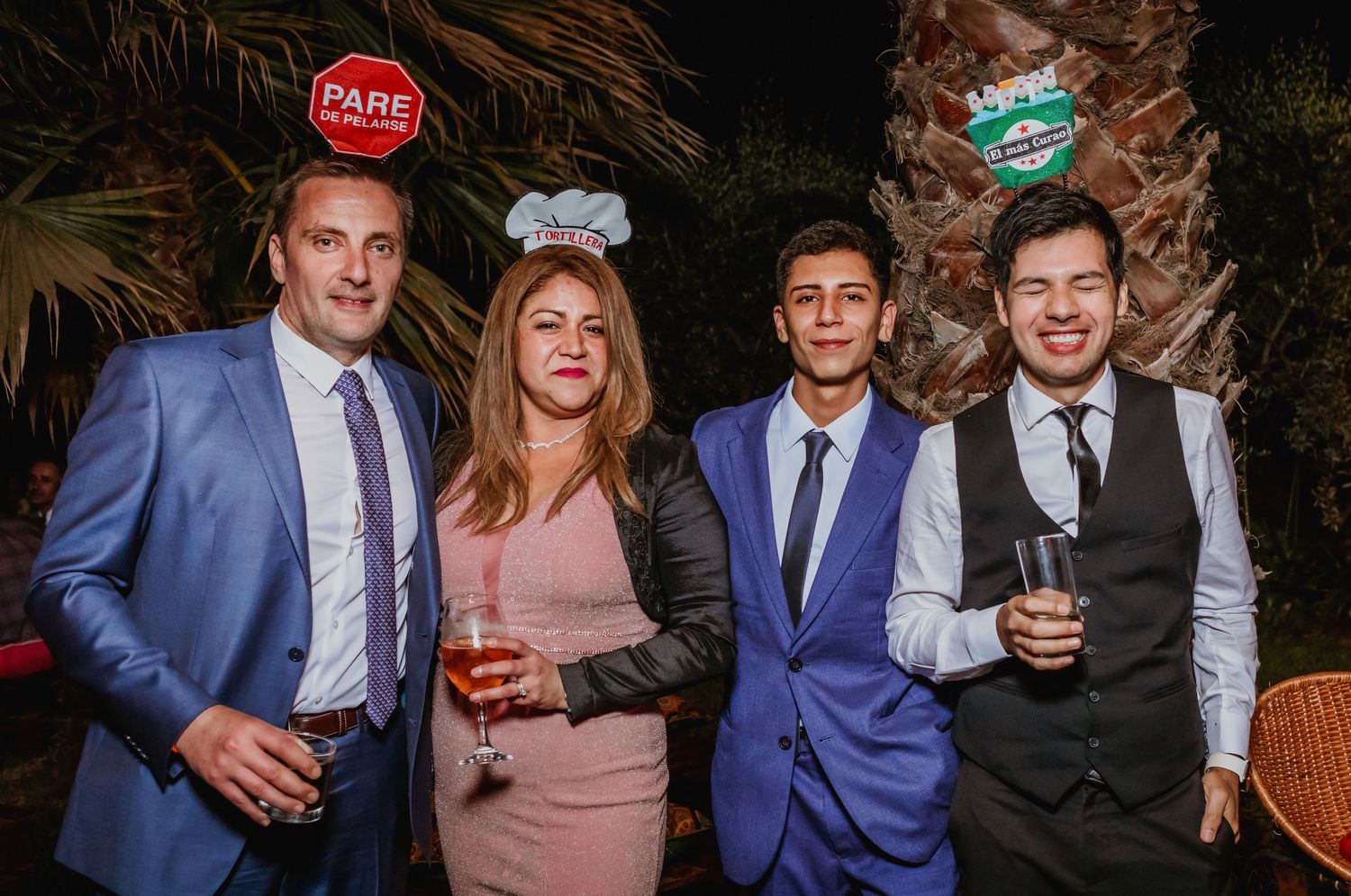 Four wedding guests pose together outdoors at night, holding drinks and wearing fun party accessories.