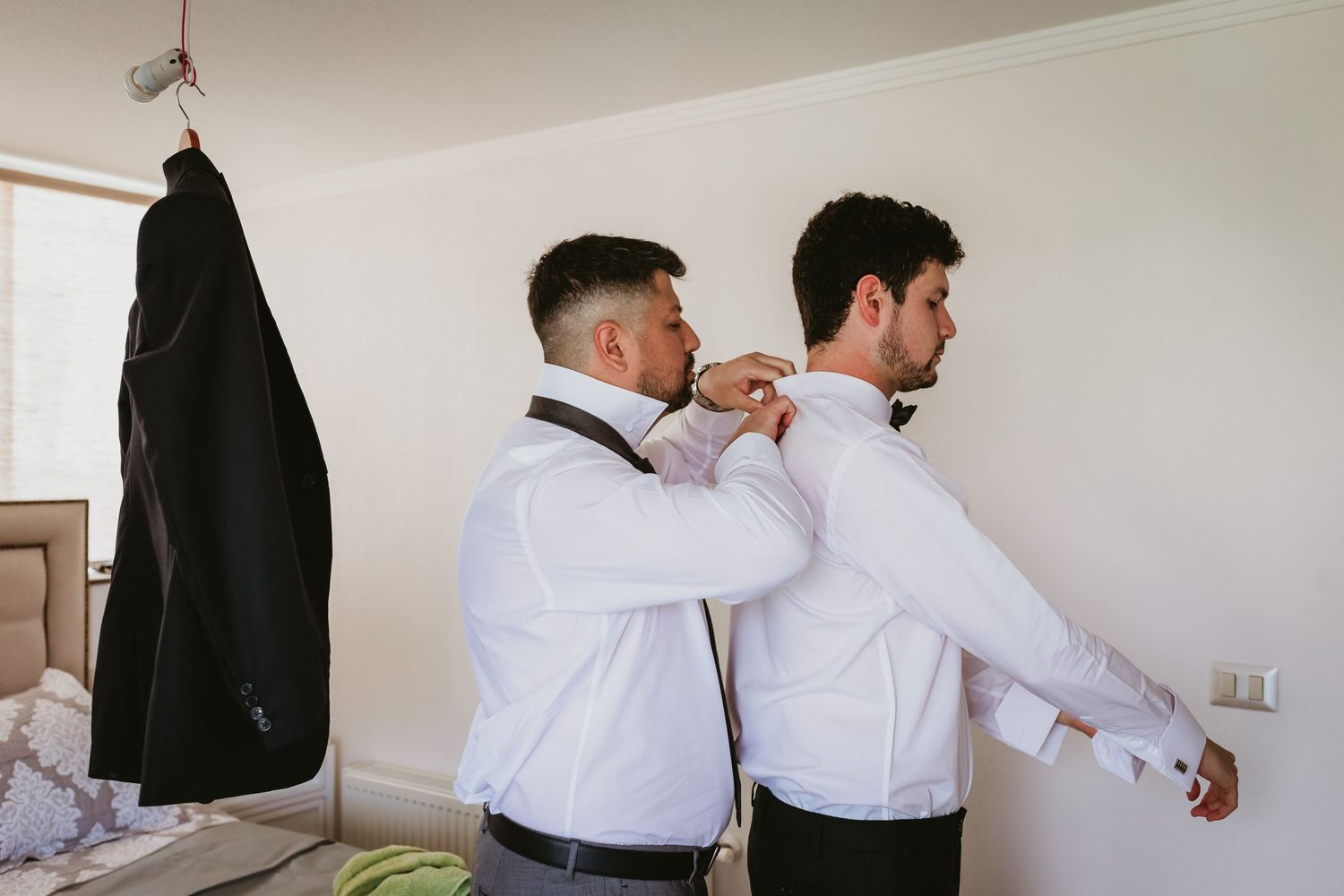 Groomsman helping groom adjust bow tie while suit hangs in background.