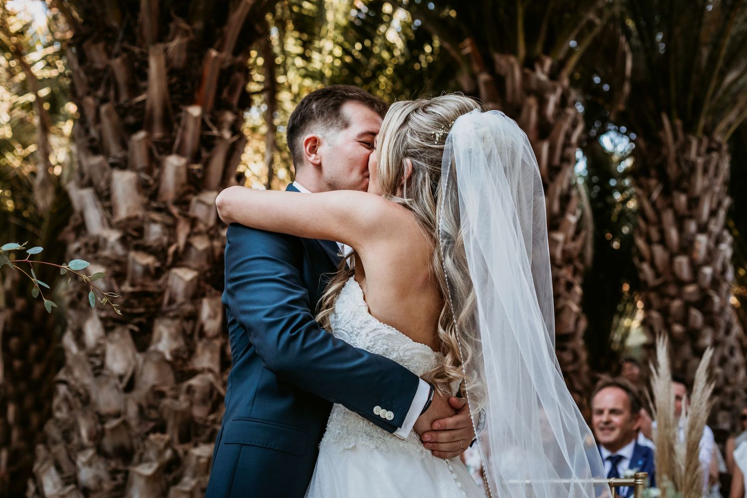 Bride and groom share their first kiss as a married couple beneath lush palm trees outdoors.