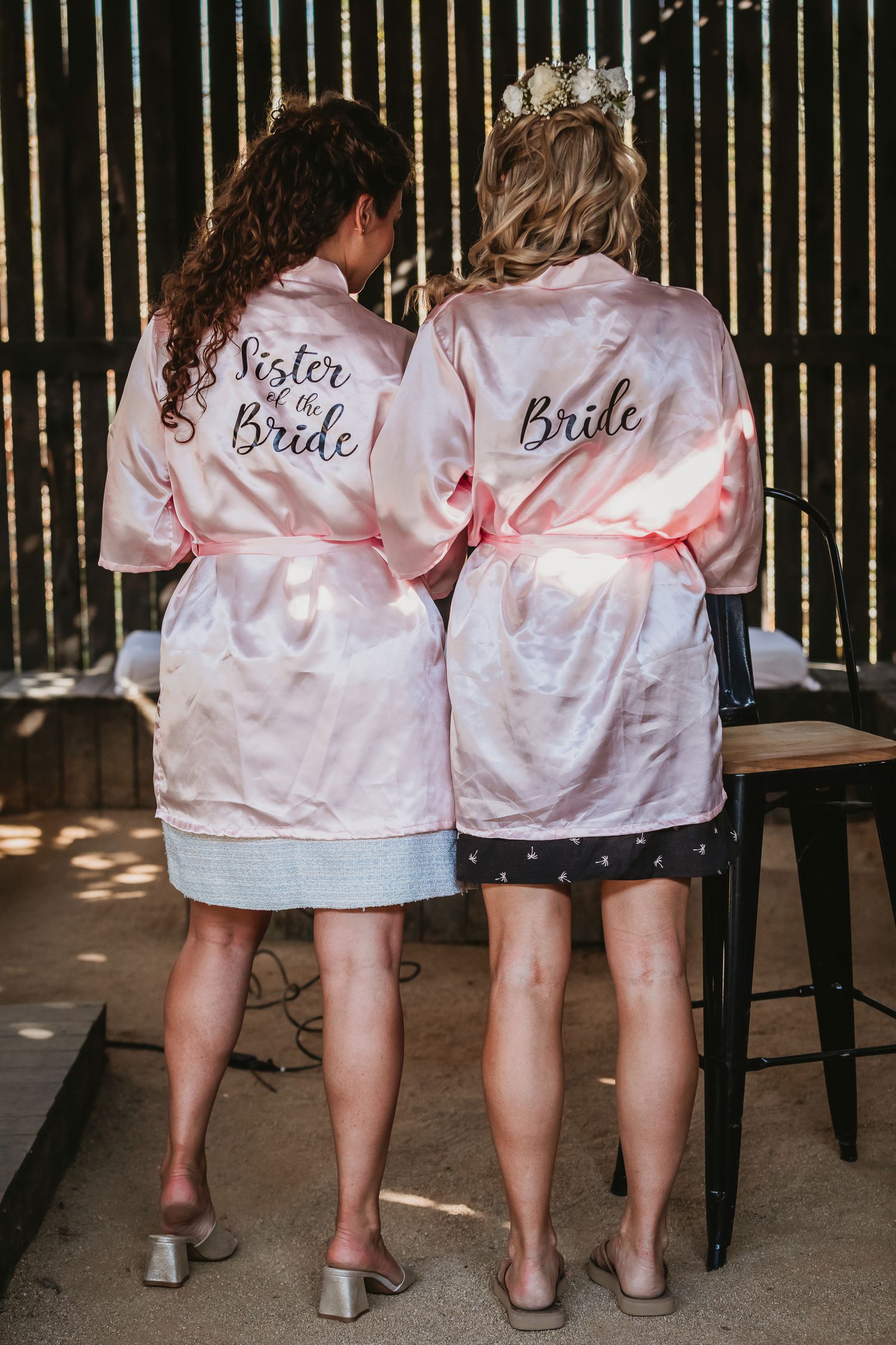 Bride and sister wearing matching pink satin robes with embroidered text at wedding.