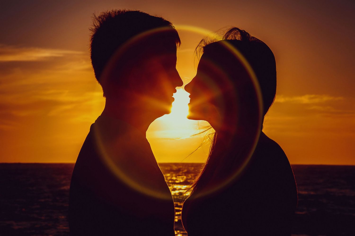 Romantic silhouette of a couple about to kiss backlit by a glowing golden ocean sunset.