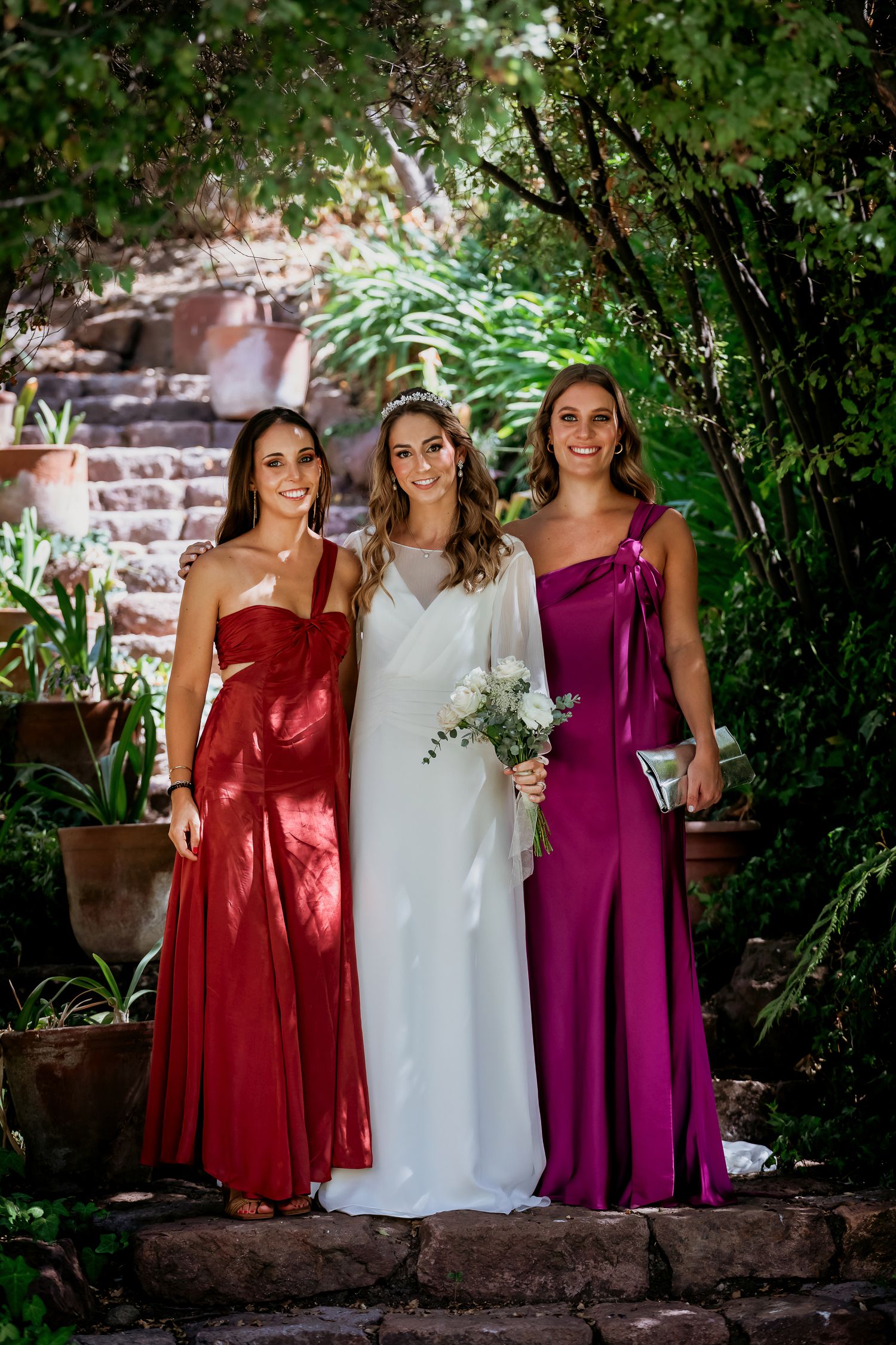 Bride in white gown poses with two bridesmaids in red and purple dresses in a lush garden.