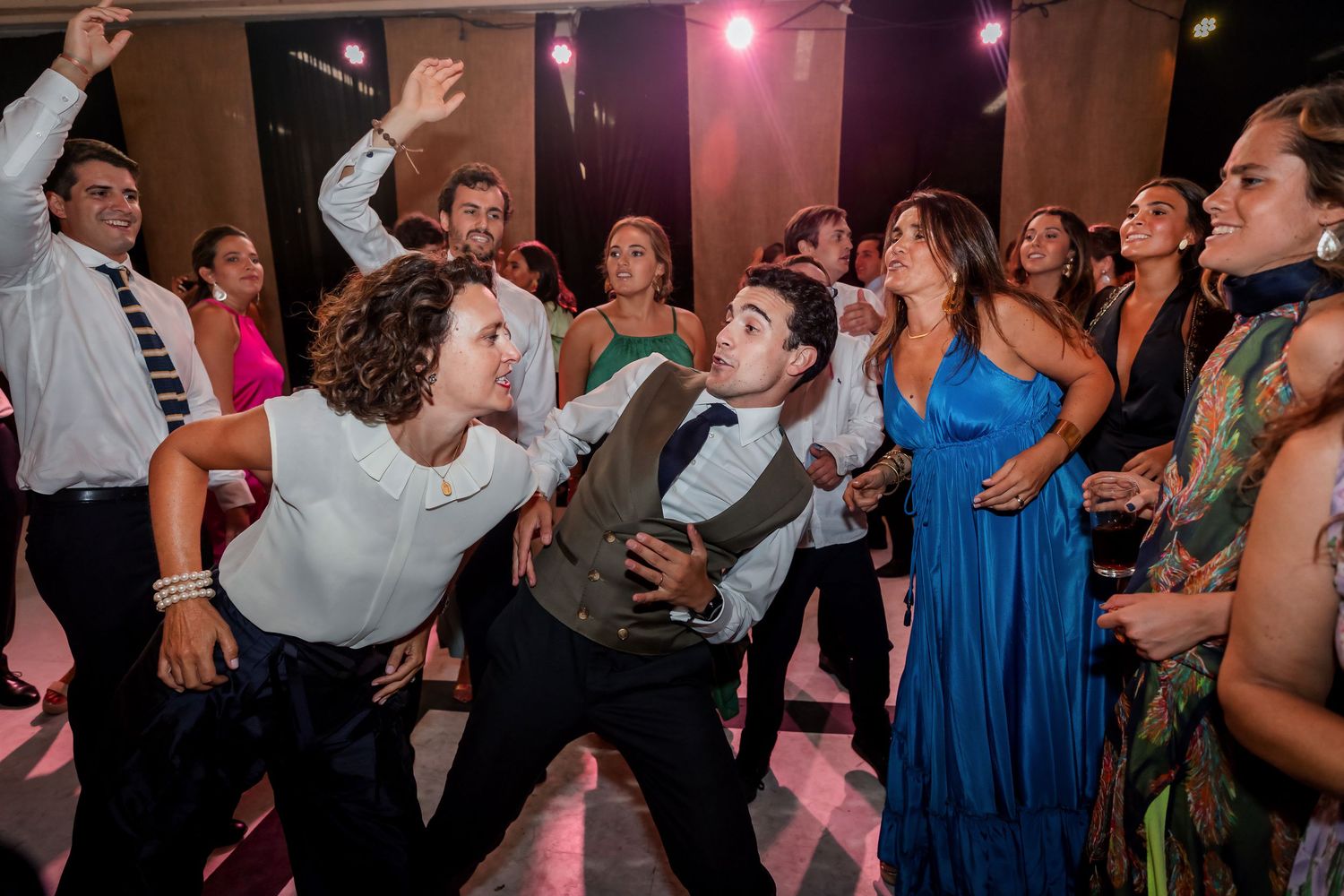 Wedding guests dancing and leaning in energetically on a colorful pink-lit reception dance floor.