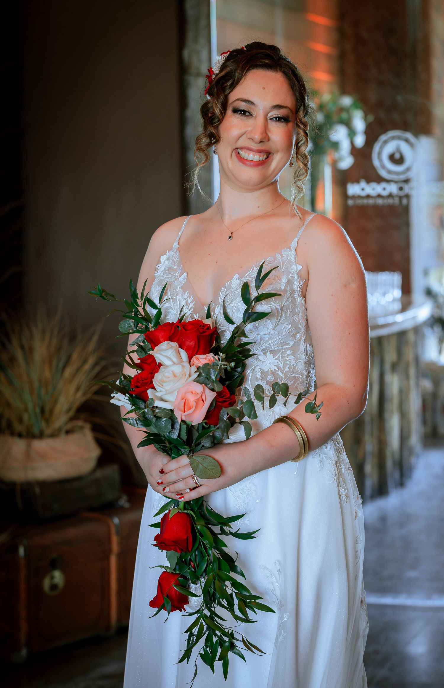 Smiling bride in white lace gown holding cascading red and pink rose bouquet with greenery at wedding venue.