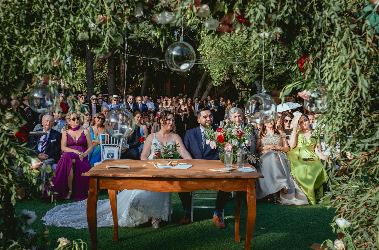 Bride and groom sit at wooden table during lush garden wedding ceremony with guests behind them.