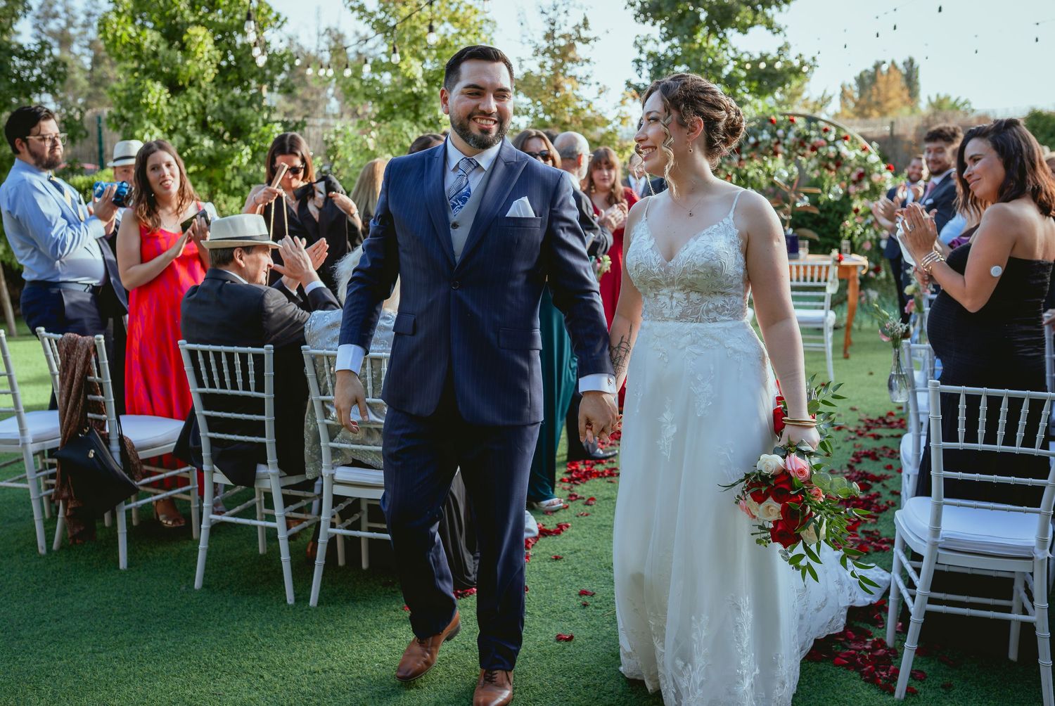 Newlyweds walk hand-in-hand down aisle as guests celebrate with confetti at outdoor garden wedding.