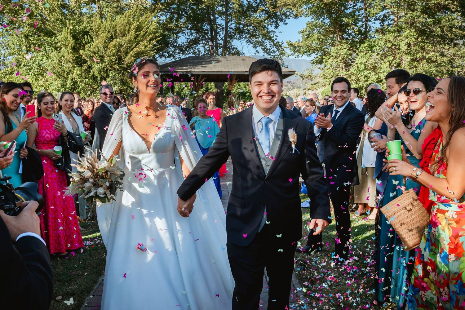 Newlyweds walking through confetti shower with smiling wedding guests celebrating outdoors.
