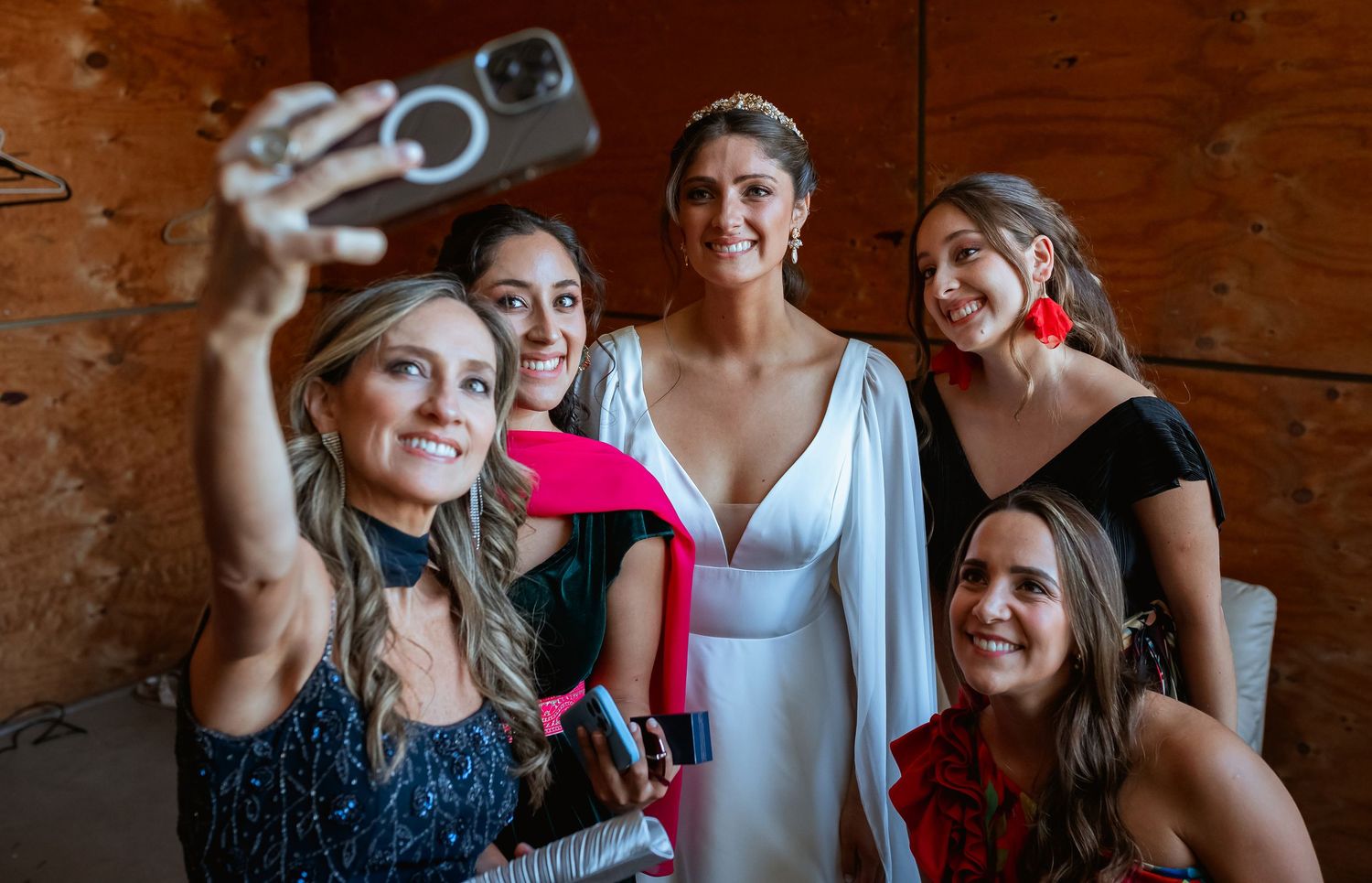 Bride in white gown posing for a selfie with smiling bridesmaids in colorful dresses before the wedding.
