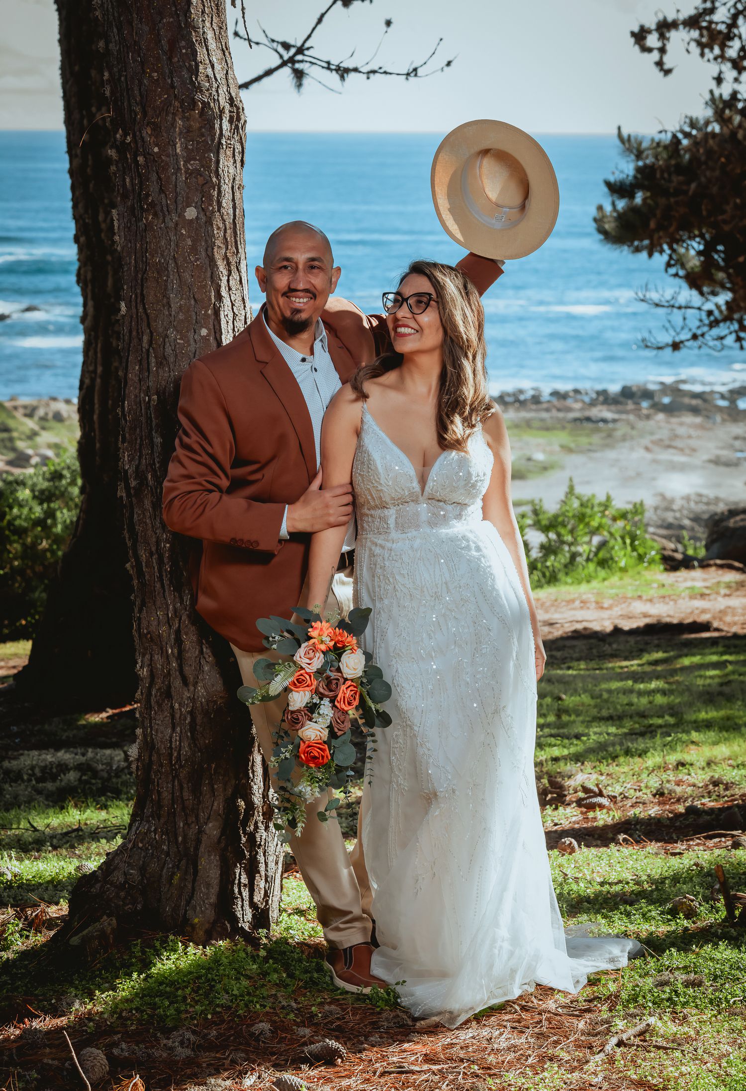 Smiling bride in a beaded gown with an orange bouquet poses beside a groom in a rust blazer holding a hat by a tree near the ocean.