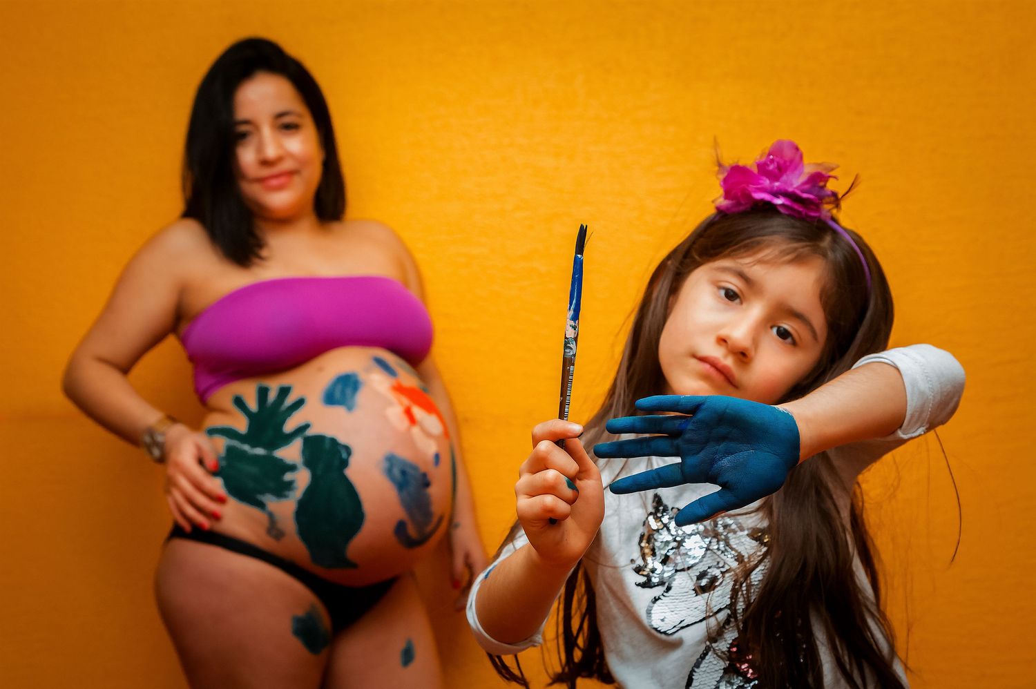 Pregnant woman and young girl artist holding a paintbrush with blue-painted hands during a fun maternity photo shoot.