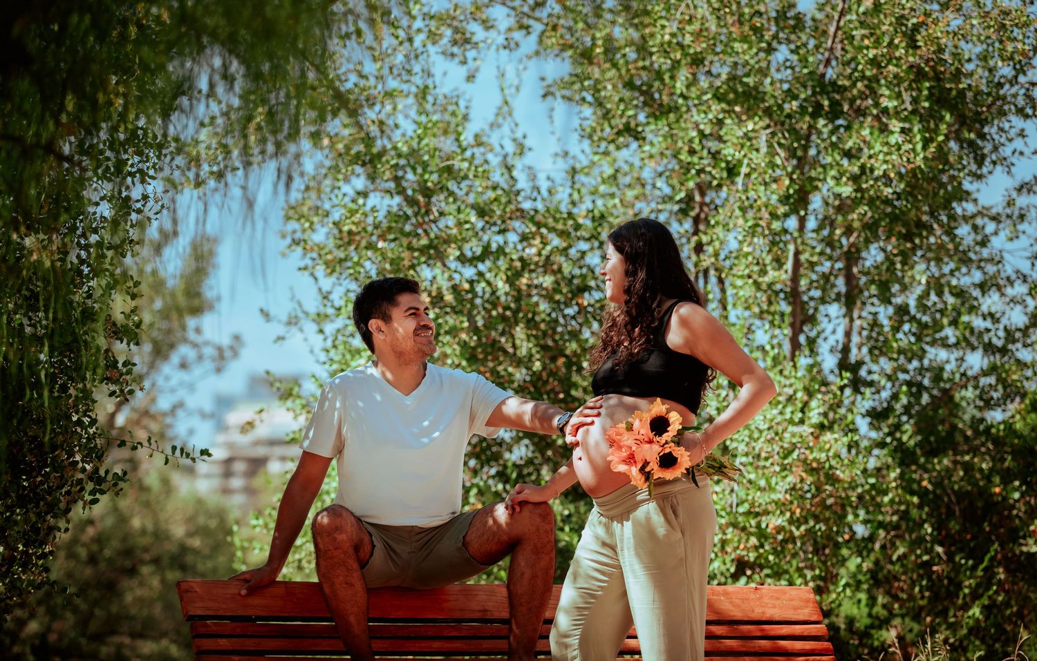 Couple sharing a tender moment on a red bench surrounded by green trees in a sunny park.