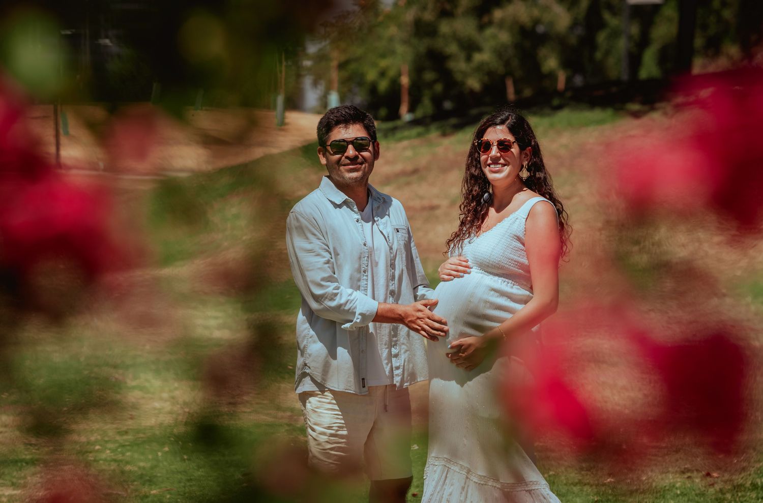 Smiling expecting couple holding baby bump surrounded by vibrant red roses in a sunny park setting.