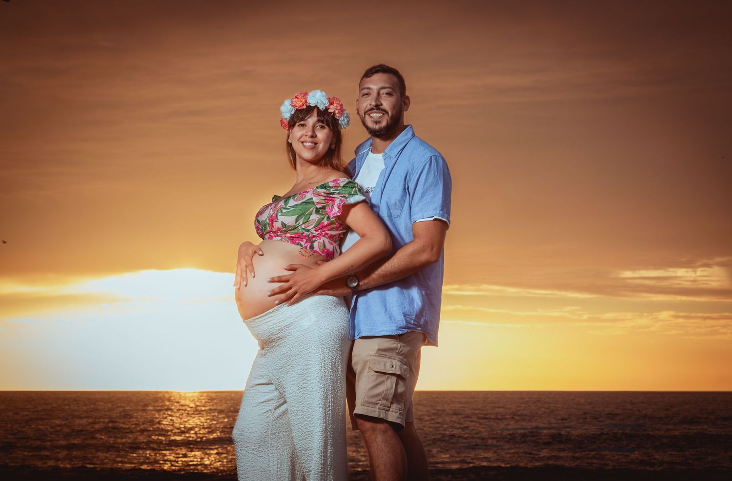 Expecting couple embraces tenderly at a stunning golden ocean sunset maternity photoshoot.