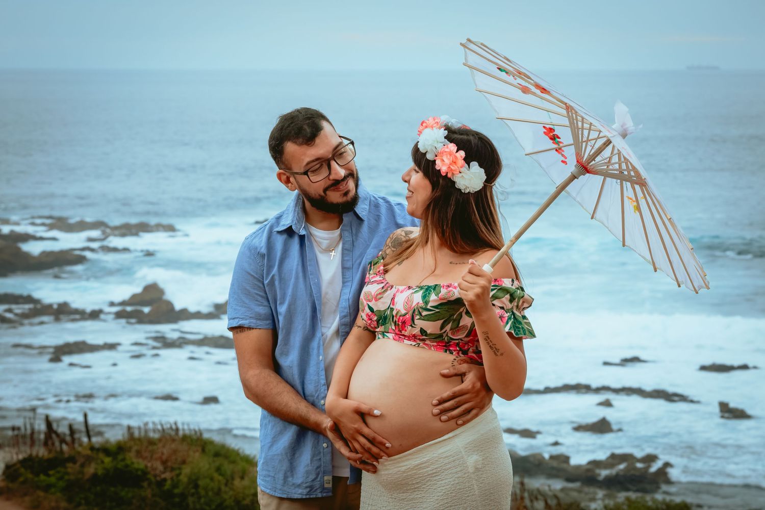 Couple embracing pregnant belly with parasol by rocky ocean shore.