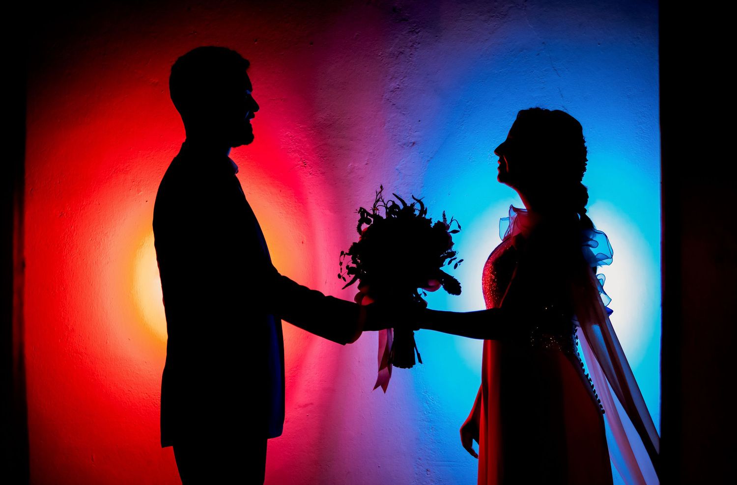 Silhouette of bride and groom exchanging flowers against a vivid red and blue gradient background.