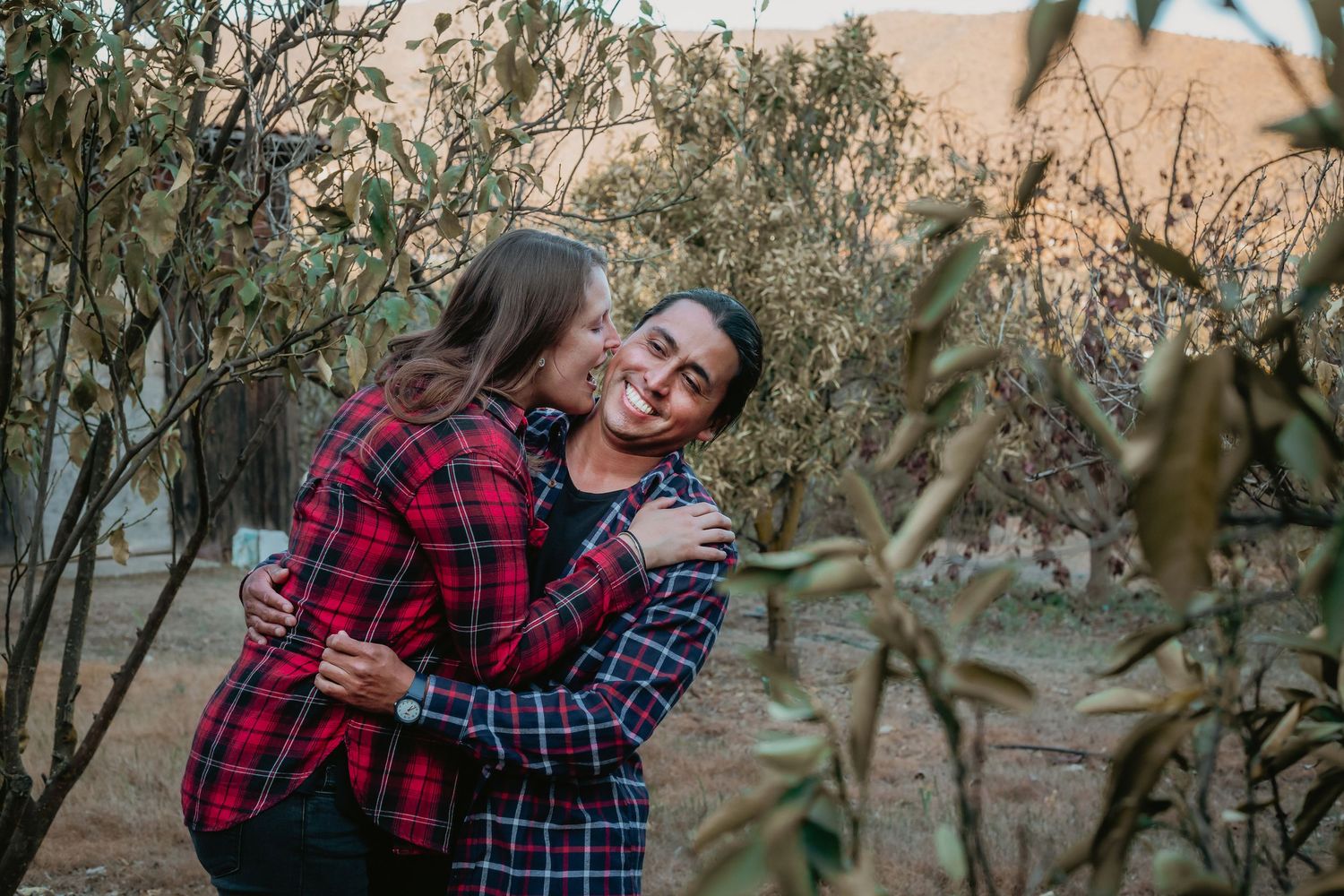 A woman kisses a man on the cheek while hugging him tightly among leafy trees outdoors.