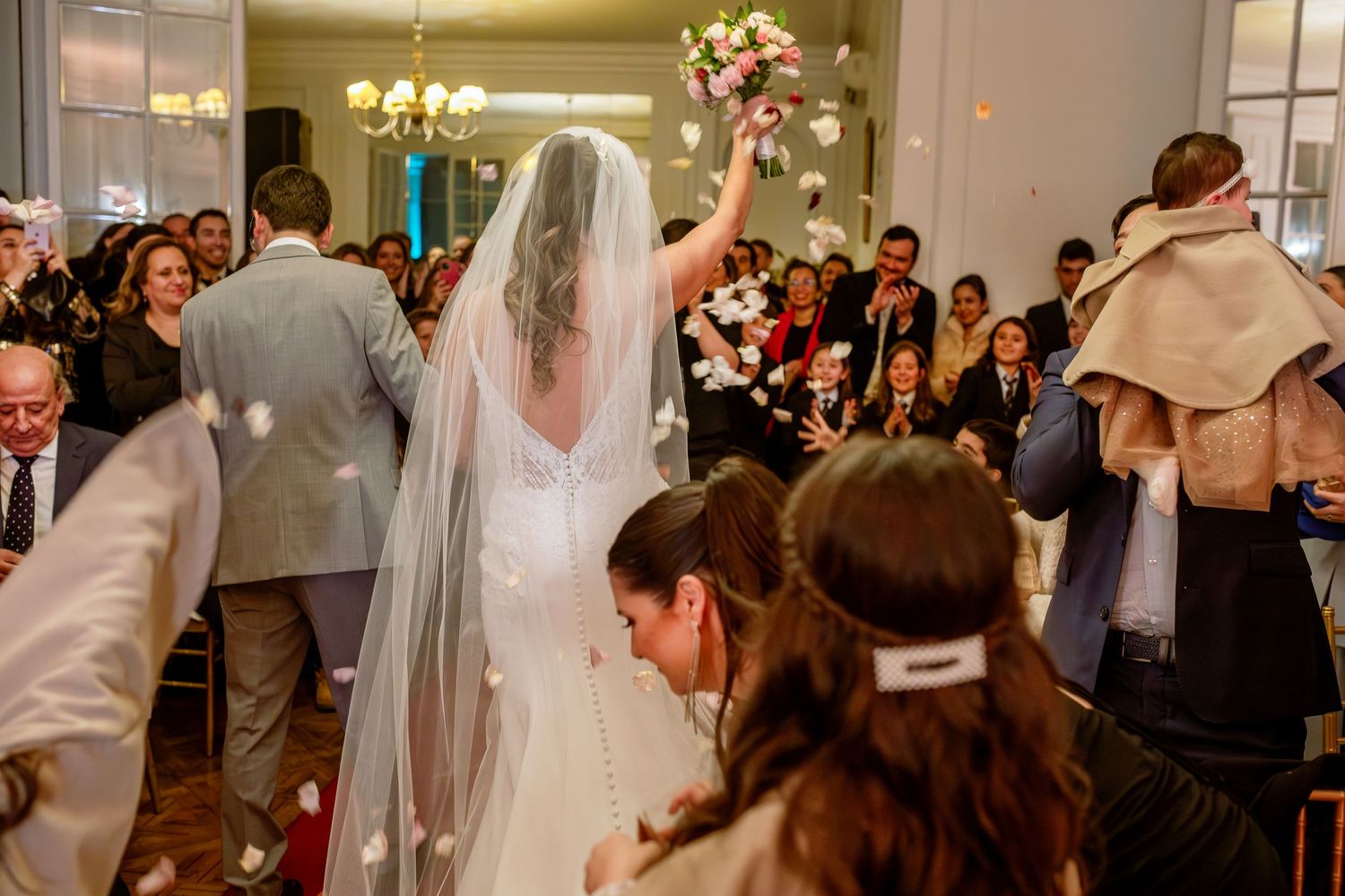 Bride walks down aisle holding bouquet aloft, surrounded by cheering guests throwing petals at indoor wedding ceremony.
