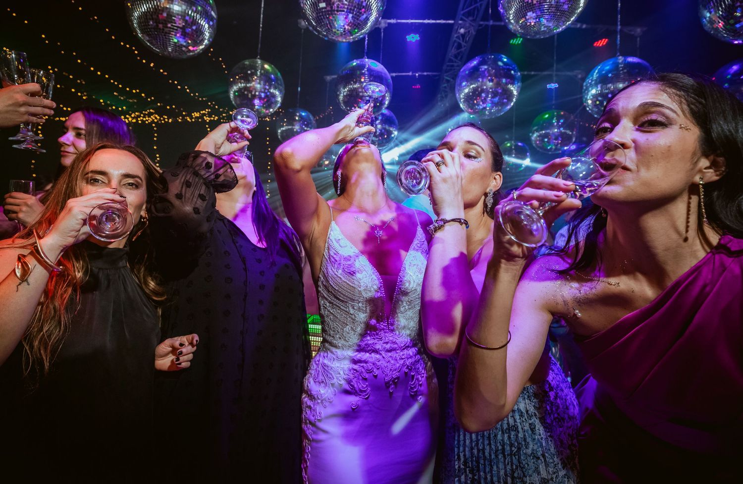 Women in elegant dresses celebrate at a disco-themed wedding reception, drinking champagne under glittering mirror ball lights.