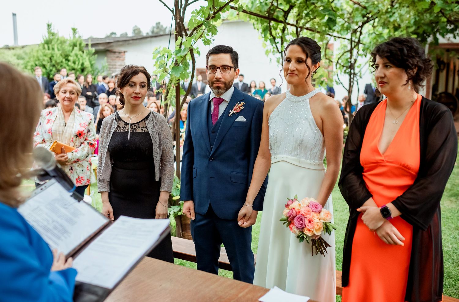 Bride and groom stand with family at outdoor wedding ceremony while officiant reads from document at table.