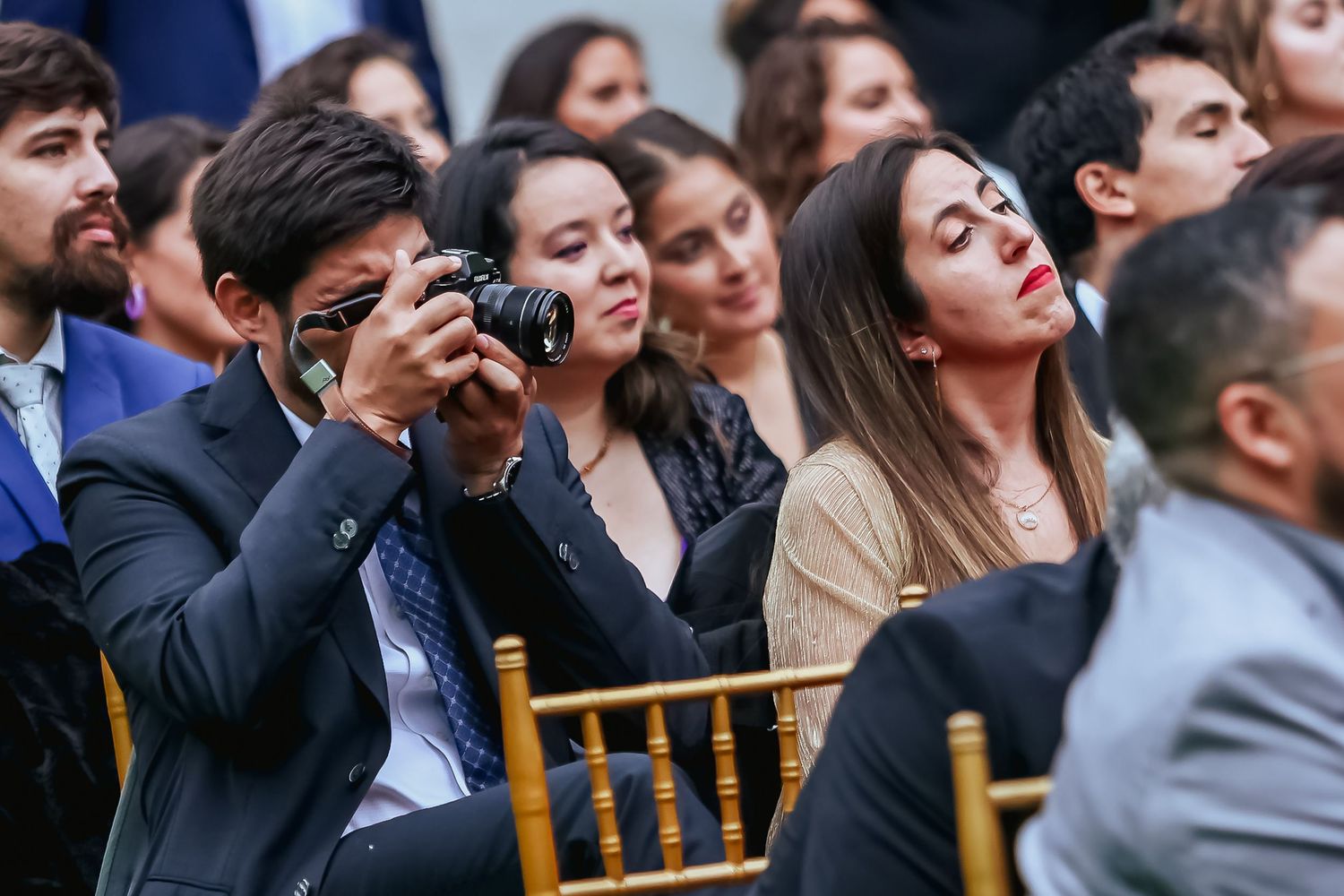 Wedding guest photographs ceremony with DSLR camera while seated among smiling guests at outdoor event.