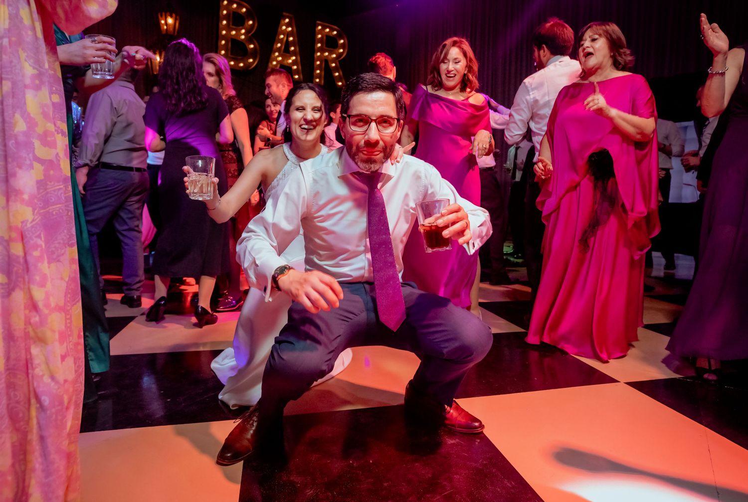 Groom in pink tie crouches playfully on dance floor while guests celebrate at lively wedding reception.