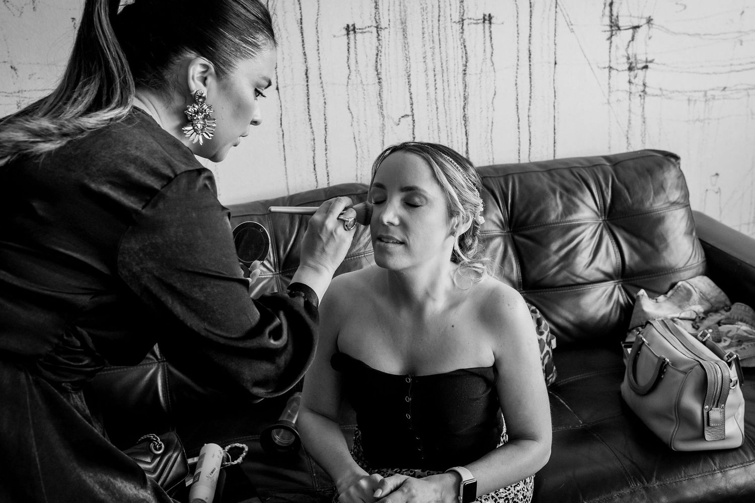Makeup artist applying cosmetics to a bride on a leather couch in black and white.